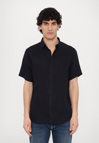 Camicia nera a maniche corte con bottoni, realizzata in tessuto leggero, con colletto classico e orlo dritto. Indossata con jeans blu.