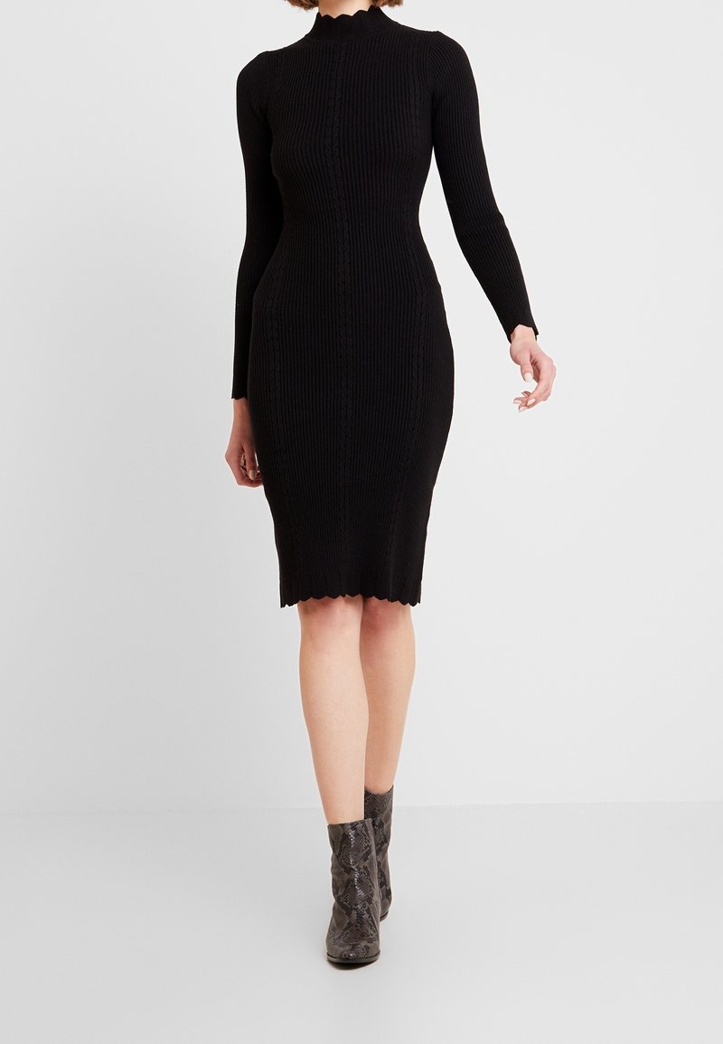 Femme portant une robe longue en maille côtelée noire à manches longues avec des bords festonnés et des bottines grises à imprimé serpent, debout sur un fond uni.