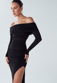 Melna kleita bez pleciem ar garām piedurknēm, savilkta dizaina, ar sānu šķēlumu, izgatavota no mīksta auduma. Zelta auskari piešķir kontrastējošu detaļu.