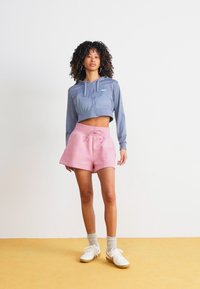 Sudadera azul ajustada con cordones, combinada con pantalones cortos rosas de cintura alta. La modelo lleva zapatillas blancas y calcetines grises, de pie sobre un suelo amarillo.
