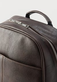 Καφέ δερμάτινο σακίδιο με υφή, που διαθέτει ασημένιο φερμουάρ και χερούλι στην κορυφή. Διακοσμημένο με ραφές κατά μήκος της μπροστινής τσέπης.