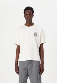 Camiseta de manga corta color crema con gráfico floral negro en el lado izquierdo del pecho, con cuello redondo y estilo relajado, hecha de material suave.