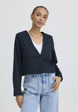 Žena oblečená v čiernom saku s výstrihom do V a jedným gombíkom, bielou košeľou, svetlomodrými džínami s vysokým pásom, zlatými náušnicami a náhrdelníkom, pózujúca s rukou v vrecku.