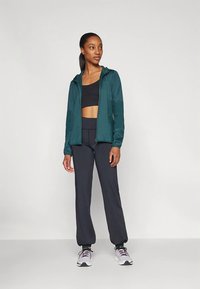 Giacca teal con zip sopra un top corto nero, abbinata a pantaloni neri e sneakers bianche. La giacca ha un cappuccio e tasche laterali.