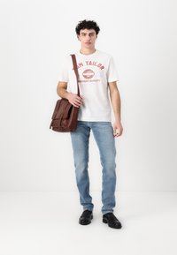 Brun lærveske med to frontlommer, hvit t-skjorte med rød grafisk tekst, lyseblå jeans og svarte sko.