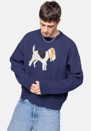 Suéter de punto azul marino con un diseño colorido de perro en el frente, con puños y dobladillo acanalados. Modelo vistiendo jeans de color azul claro.
