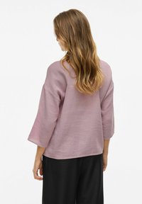 Světle růžová blůza s tříčtvrtečními rukávy, měkkou texturou a volným střihem, kombinovaná s černými kalhotami. Čistý design, žádné viditelné vzory.