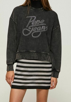 Femme portant un sweat-shirt noir délavé à manches longues avec un logo « Pepe Jeans » en strass et une mini-jupe tricotée rayée noir et blanc.