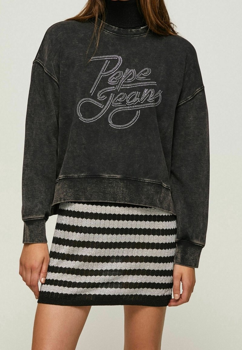 Femme portant un sweat-shirt noir délavé à manches longues avec un logo « Pepe Jeans » en strass et une mini-jupe tricotée rayée noir et blanc.