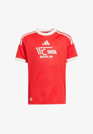 Garçon en tenue de football rouge du 1. FC Union Berlin, en train de frapper le ballon, portant un maillot rouge, un short blanc, des chaussettes rouges et des crampons noirs, sur un fond uni.