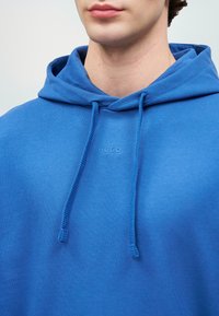 Φούτερ με κουκούλα σε έντονο μπλε, κατασκευασμένο από μαλακό ύφασμα. Διαθέτει λογότυπο μπροστά, κορδόνι και άνετη εφαρμογή. Λείο υφασμά, χωρίς σχέδια.