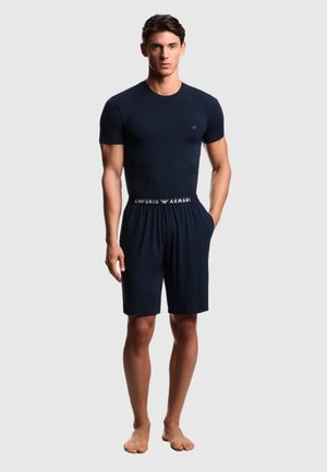 Uomo in piedi a piedi nudi che indossa una maglietta nera aderente e pantaloncini neri con fascia in vita "Emporio Armani" su sfondo grigio.