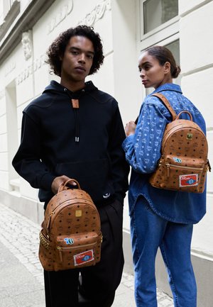 Zaino in pelle marrone con motivo in rilievo, finiture dorate e una tasca verticale con zip sul fronte. Indossato da una persona con una giacca di denim. Felpa nera nelle vicinanze.