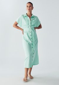 Mėtų žalios spalvos mygtukinė midi suknelė su trumpomis rankovėmis, dviem krūtinės kišenėmis, diržu liemeniui ir auksiniais mygtukais. Lygi, lengva medžiaga.