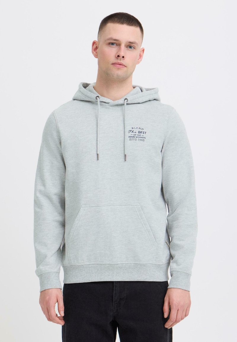 Jeune homme portant un sweat à capuche gris clair avec poche avant et un pantalon noir, debout devant un fond blanc uni.