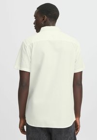 Camisa de manga corta en un tono crema claro, de tela texturizada con cuello tipo solapa y un patrón sutil. La vista trasera muestra detalles de costura y un dobladillo curvado.