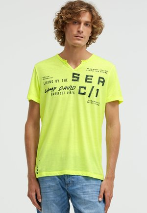 Junger Mann mit lockigem Haar, der ein neongelbes V-Ausschnitt-T-Shirt mit schwarzem Aufdruck und blaue Jeans trägt und vor einem schlichten Hintergrund steht.