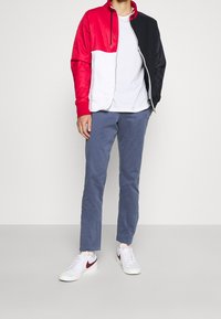 Červená, bílá a černá zipová bunda; šedé bavlněné kalhoty; bílé tenisky s červenými detaily. Hladký materiál, uvolněný střih a ležérní styl.