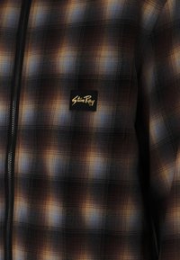 Plaid zip-up jacket featuring dark brown, blue, and orange tones with a branded black patch. Smooth texture with checkered pattern.