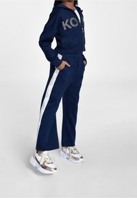 Námořnický modrý teplákový set s cropped hoodie zdobeným kamínkovým logem "KORS" a odpovídajícími kalhotami s bílými bočními pruhy. Masivní zlaté tenisky.