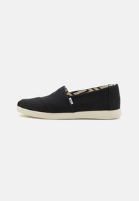 Schwarze Leinenschlupfschuhe mit strukturierter Oberfläche, weißer Gummisohle und gemusterter Fütterung, die am Öffnungsrand sichtbar ist. Keine Schnürsenkel oder Verschlüsse.