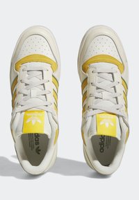 Zapatillas blancas con acentos de ante amarillo, con puntera perforada, tres rayas verdes y cordones texturizados. Etiqueta de logo amarilla en el talón.