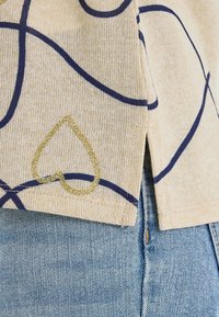 Top beige con líneas azul marino y un detalle de corazón dorado con brillo, que presenta una textura suave y un dobladillo dividido. Combinado con denim azul claro.