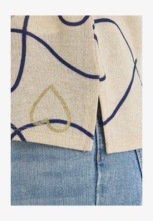 Top beige con líneas azul marino y un detalle de corazón dorado con brillo, que presenta una textura suave y un dobladillo dividido. Combinado con denim azul claro.