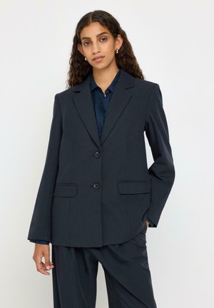 Ung kvinde med langt krøllet hår iført en mørk marineblå to-knap blazer og matchende bukser, stående mod en ensfarvet lys baggrund.
