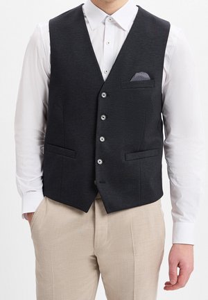 Homme portant un gilet noir boutonné sur une chemise blanche à manches longues et un pantalon beige, les mains dans les poches.