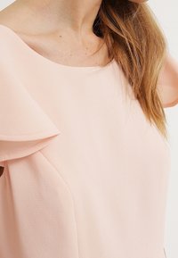 Svetle růžové šaty s kulatým výstřihem a volánovými rukávy. Materiál má texturovaný povrch, což dodává designu hloubku.