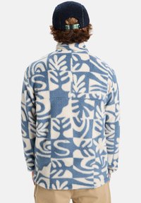 Fleecejakke i krem og blå med blomsterdesign, høy krage, lange ermer og løs passform. Utsmykket med en blå caps.