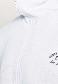 Sweat à capuche zippé gris clair en mélange de coton doux, avec un logo brodé en bleu marine et un col montant avec une fermeture éclair complète.