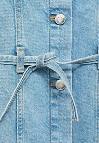 Jasnoniebieska tkanina dżinsowa z przewiązanym paskiem wloopach na pasek oraz srebrnymi guzikami z wytłoczonym napisem "MIG DENIM".