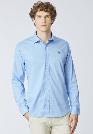 Giovane uomo con capelli ricci che indossa una camicia azzurra con bottoni e un piccolo logo sul petto, pantaloni beige, con le mani nelle tasche.