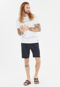 Miestna biela košeľa s krátkym rukávom s horizontálnymi multicolor pruhmi, v kombinácii s námorníckymi kraťasmi a hnedými sandálmi. Viditeľné tetovanie na rukách.