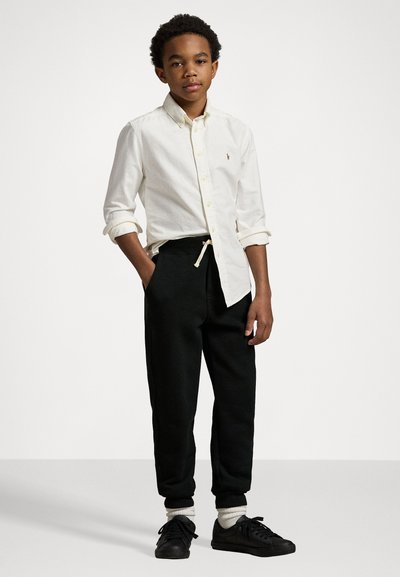Chemise blanche à manches longues avec boutons, pantalon de survêtement noir avec cordon de serrage, baskets noires et chaussettes blanches mi-mollet. Fond uni.