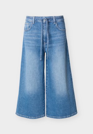 Culottes larges en denim bleu moyen avec poches avant, passants pour ceinture et ceinture à nouer sur un fond clair.