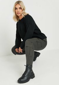 Suéter negro de punto acanalado, pantalones cargo oliva con bolsillos laterales y botines negros de cordones con suelas gruesas; modelo en cuclillas.