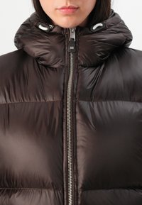 Giubbotto imbottito marrone con colletto alto, chiusura a zip e dettagli con cordini; presenta una texture liscia e lucida e un design trapuntato.