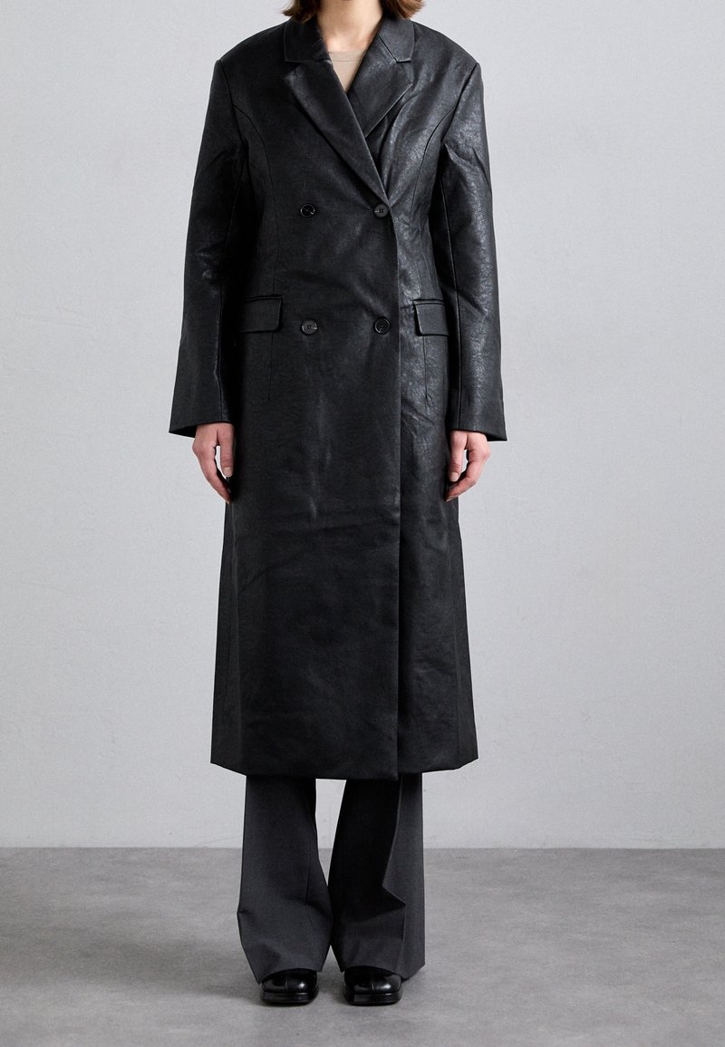 Personne portant un long manteau en cuir noir croisé avec de larges revers, un pantalon gris et des chaussures noires sur un fond gris uni.