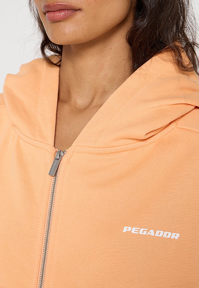 Helle orange Zip-Hoodie aus weichem Stoff. Verfügt über eine große Kapuze und ein weißes "PEGADOR"-Logo auf der Brust. Glatte Textur und minimalistisches Design.