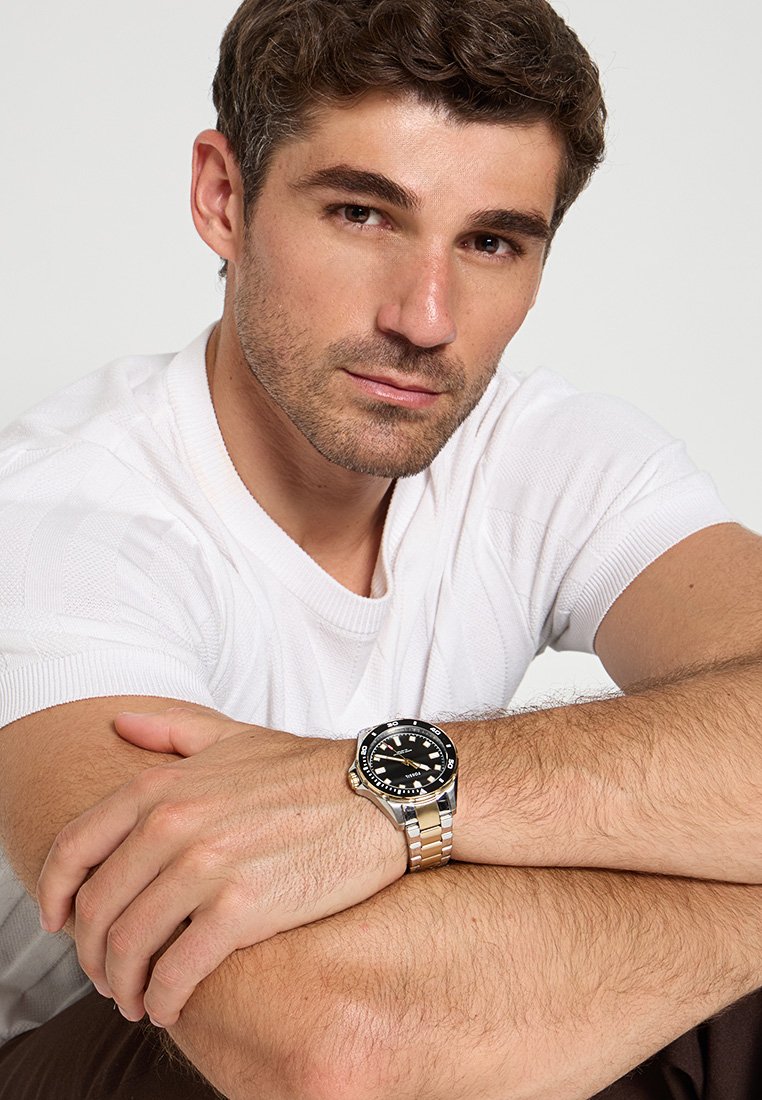 Orologio con quadrante nero e bracciale in acciaio inossidabile e tono oro, caratterizzato da una cassa rotonda, grandi markers e una lunetta girevole.