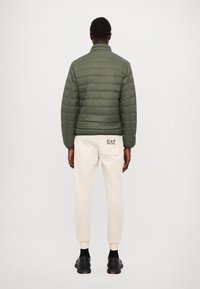 Green padded jacket with a stand collar, paired with cream sweatpants featuring a rear pocket logo. Black sneakers complete the outfit.