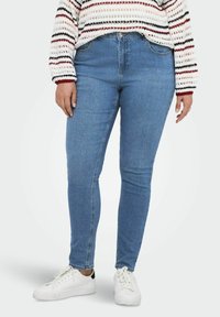 Mėlynos džinsinės siaurėjantys džinsai su vidutiniu juosmeniu, puošti subtiliais siūlais ir klasikiniu penkių kišenių dizainu, derinami su baltais sportbačiais.