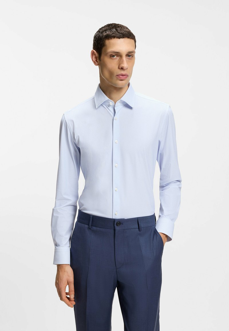 Homme portant une chemise bleu clair boutonnée et rentrée dans un pantalon bleu foncé, debout avec une main dans la poche, sur un fond uni.