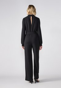 Black long-sleeve jumpsuit with a fitted waist, back keyhole cutout, and wide-leg trousers, displayed from the back. Smooth fabric texture.