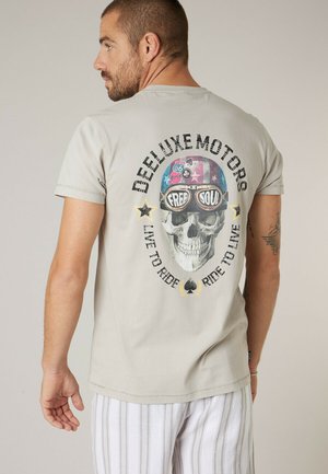 Homme portant un t-shirt beige avec un crâne coloré portant un casque et des lunettes, texte « DEELUXE MOTORS », « FREE SOUL » et « LIVE TO RIDE ».