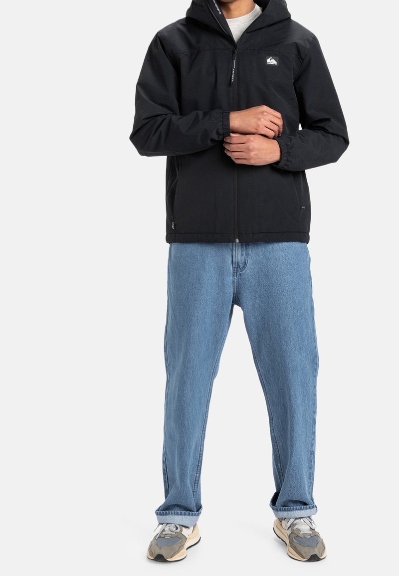 Personne portant une veste à capuche noire, un jean bleu clair ample et des baskets grises, debout les bras croisés à la taille.