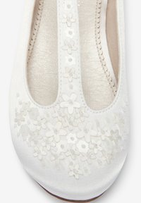 Chaussure plate en satin blanc avec une bride en T, décorée de petites appliques florales blanches et de perles sur le bout et la bride.